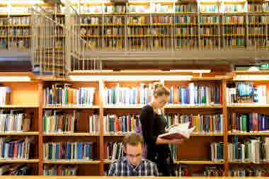 Två studenter på bibliotek. En står upp och läser i en bok, den andra sitter ner. 