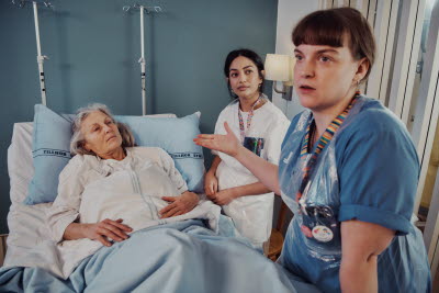 Sjuksköterska diskuterar med kollega eller anhörig kring patient som ligger i vårdsäng.