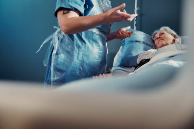 Patient i vårdsäng, sjuksköterska med plastförkläde, gestikulerar.
