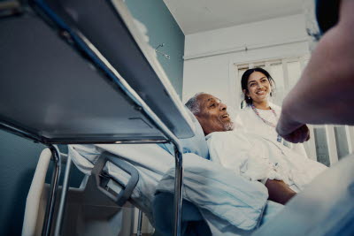 Bild tagen i grodperspektiv. Patient i vårdsäng med glad sjuksköterska bredvid.