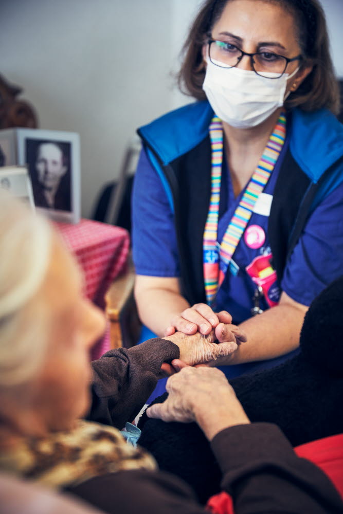 Sjuksköterska med munskydd håller en boende i handen. Omvårdnad, RIO äldreboende.