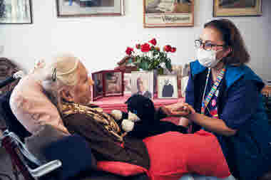 Sjuksköterska med munskydd håller en boende i handen i den boendes rum. Omvårdnad, RIO äldreboende.