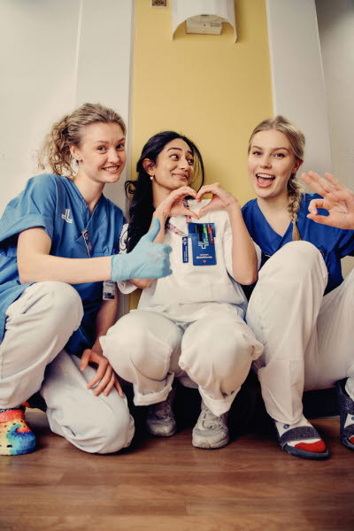 Tre leende kvinnliga kollegor sittande på huk. Handske med texten Vi fixar det! , tummen upp, hjärta och ok-tecken. Glädje, flams, gemenskap