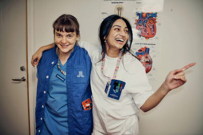 Två glad kollegor som stöttar varandra, den en skrattar och pekar mot någon utanför bild. Glädje, gemenskap, flams.