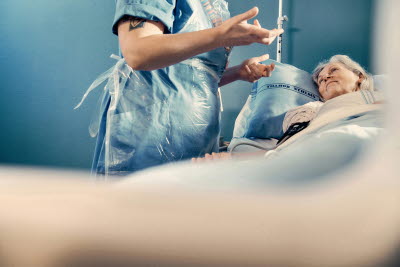 Patient i vårdsäng, sjuksköterska med plastförkläde, gestikulerar.