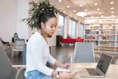 Kvinna i ett bibliotek med laptop, telefon och anteckningsblock.