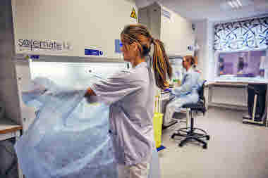 Biomedicinsk analytiker på Gävle sjukhus mars 2021. BMA sätter på sig skyddsutrustning vid dragskåp. Kollega arbetar bakom. 