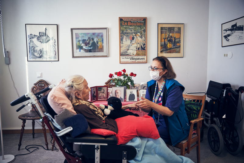 Sjuksköterska med munskydd håller en boende i handen i den boendes rum. Omvårdnad, RIO äldreboende.