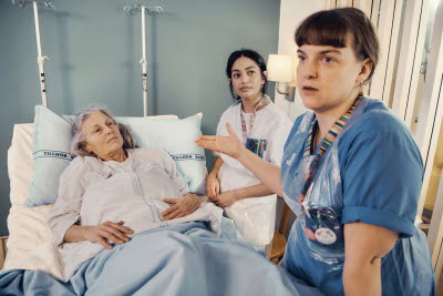 Sjuksköterska diskuterar med kollega eller anhörig kring patient som ligger i vårdsäng.