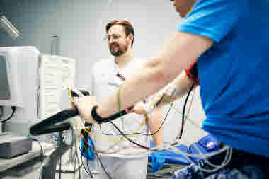 Biomedicinsk analytiker, klinisk fysiologi, arbets-EKG, leende man övervakar patient som cyklar uppkopplad elektroder, Karolinska sjukhuset i Solna