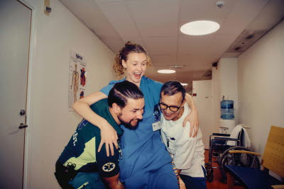 Skrattande kollegor i en korridor som busar med varandra. Två personer lyfter en tredje. Glädje, gemenskap och flams.